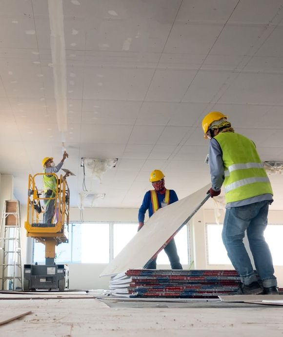 Construction workers installing drywall on a ceiling. Yellow hard hats, high-vis vests. Inside building.