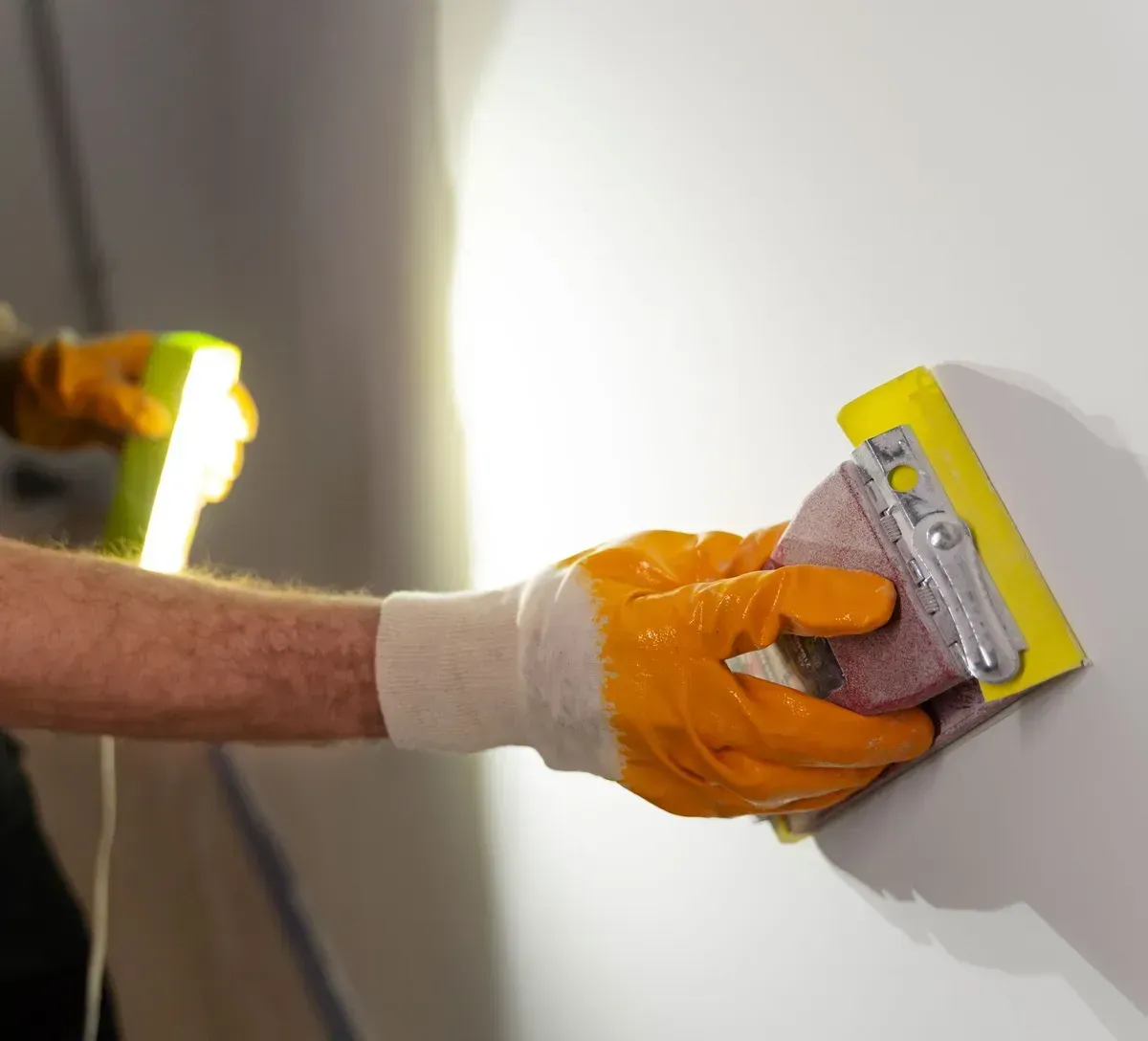 Person sanding a wall corner with a sanding block, illuminated by a work light.