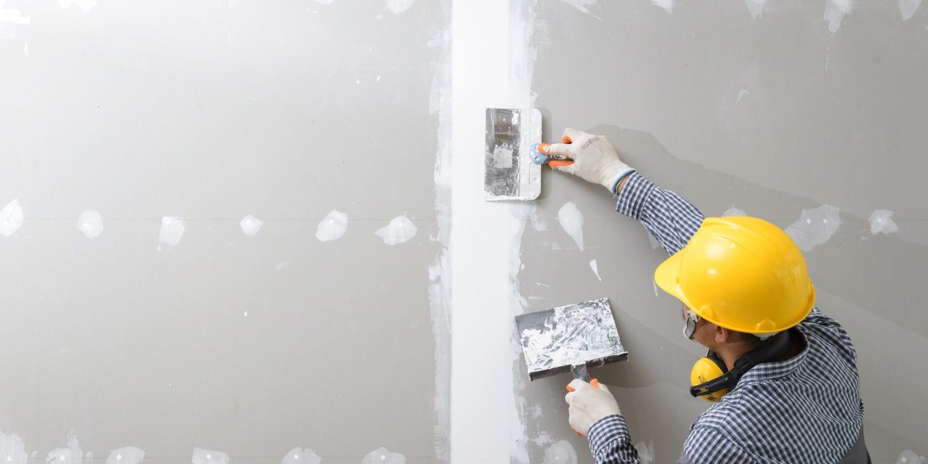 Person wearing a yellow hard hat and gloves, applying drywall mud to a wall.