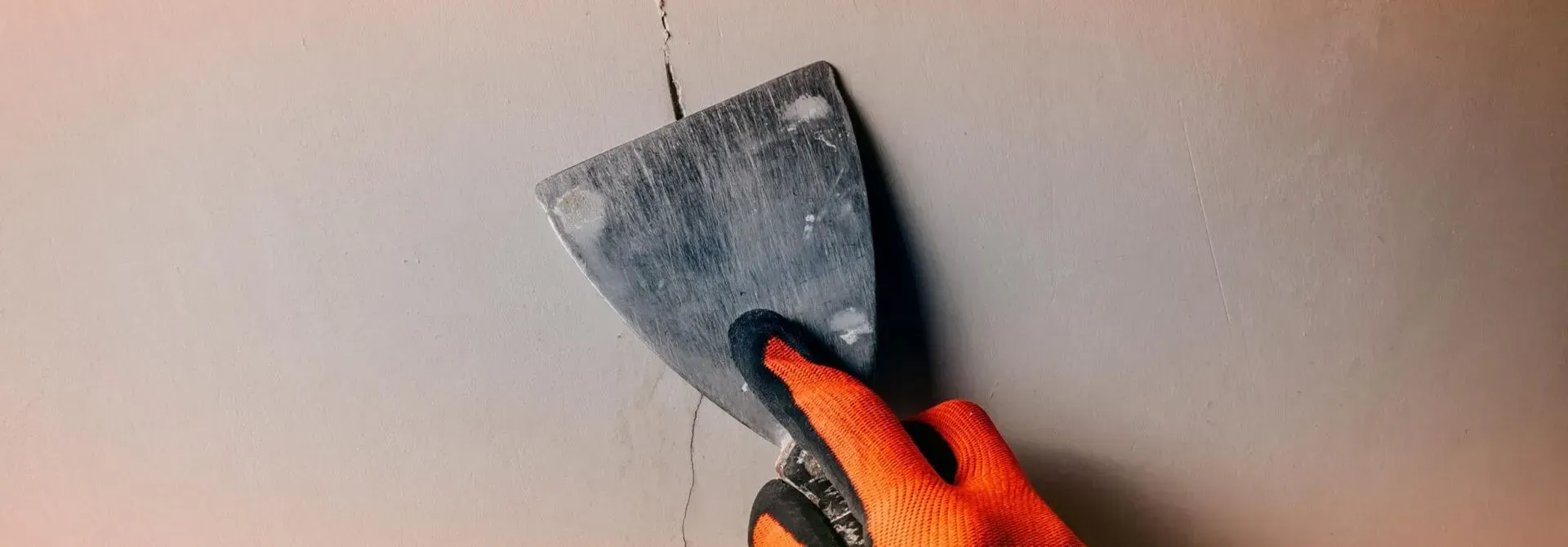 Person with orange-gloved hand using a putty knife to fill a crack in a white wall.