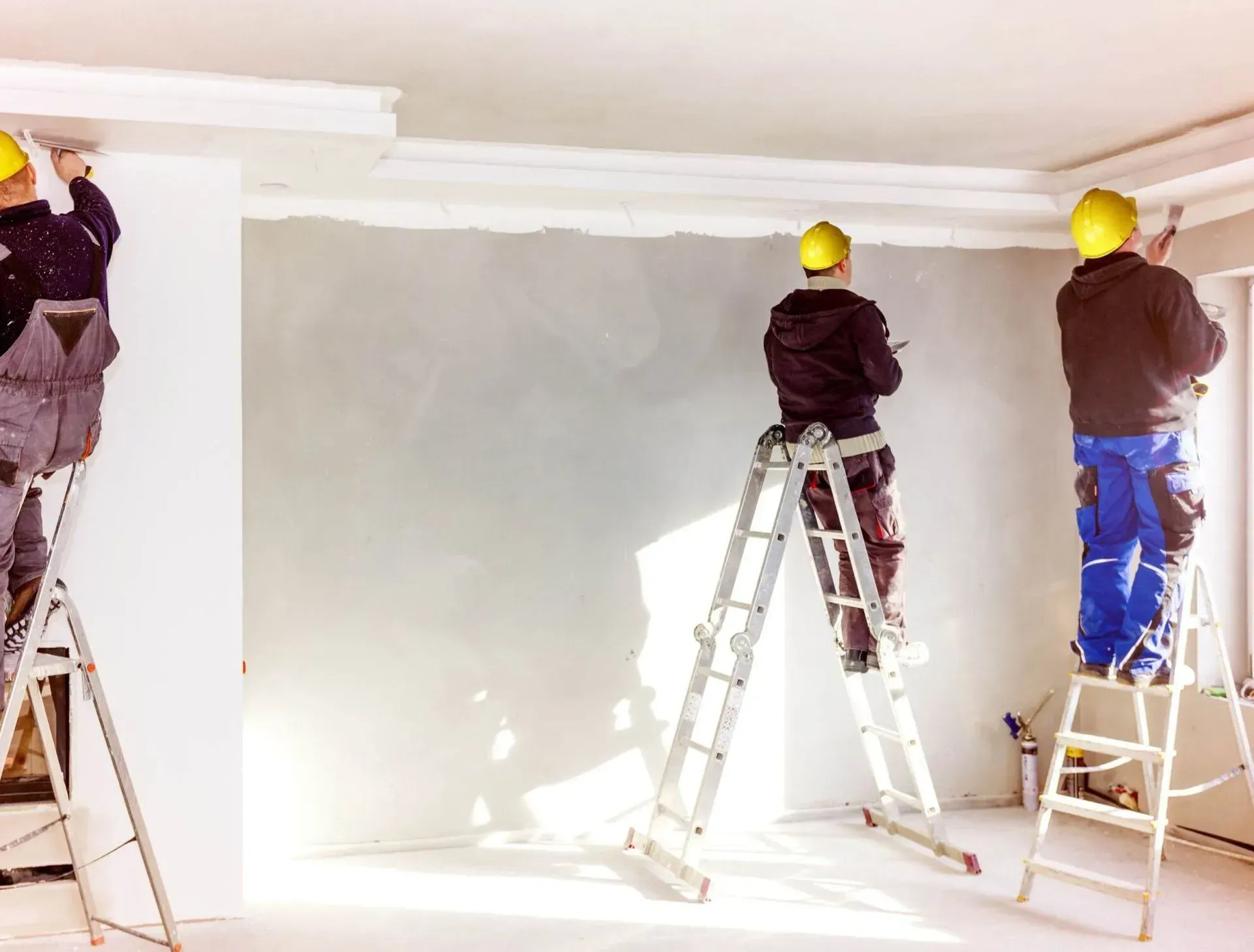 Three workers installing crown molding in a room, using ladders and wearing hard hats.