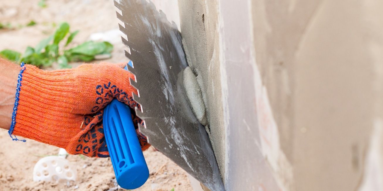Hand in orange glove using a notched trowel to apply mortar to a wall.