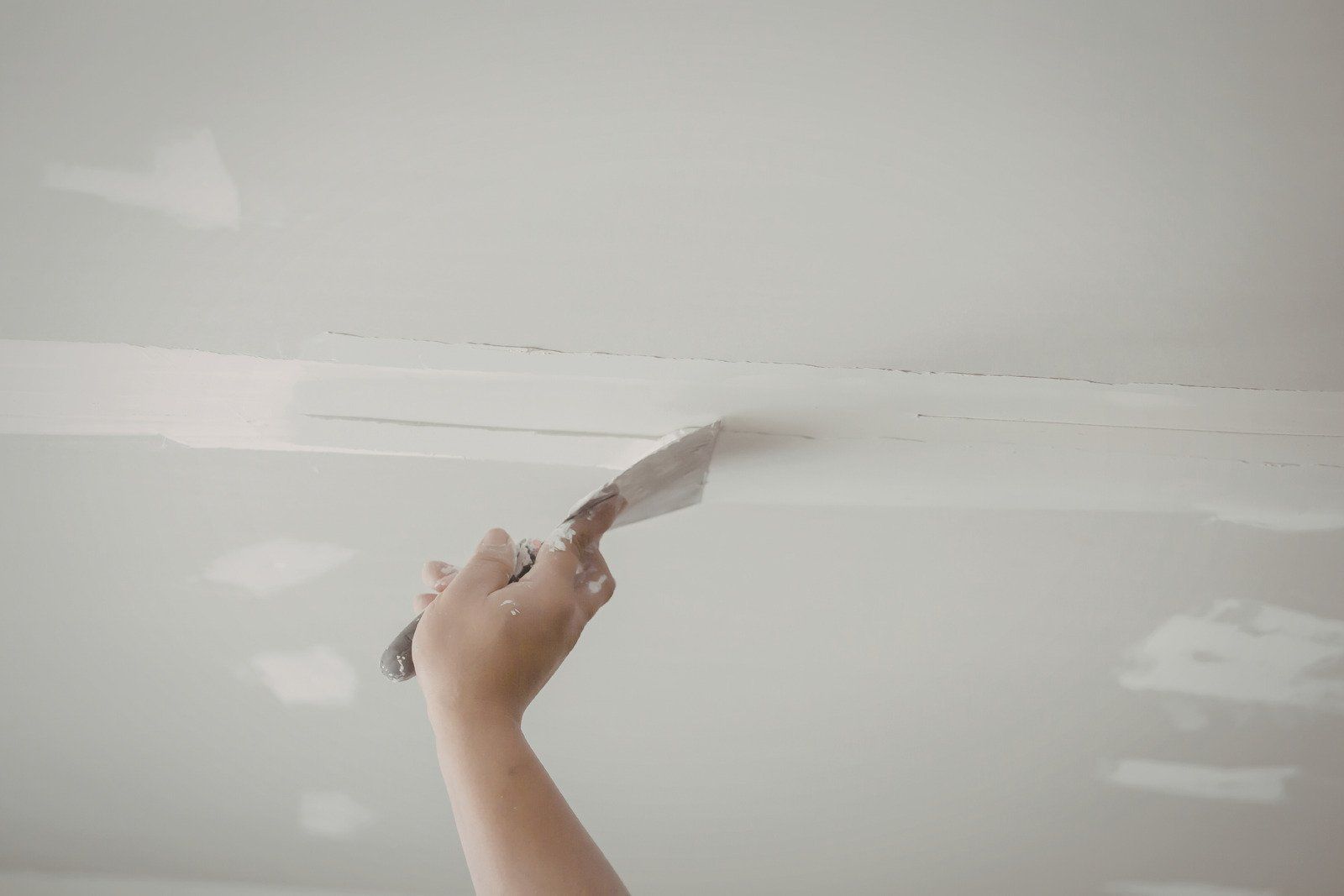 Man on a ladder using a drywall knife to apply joint compound to a ceiling.