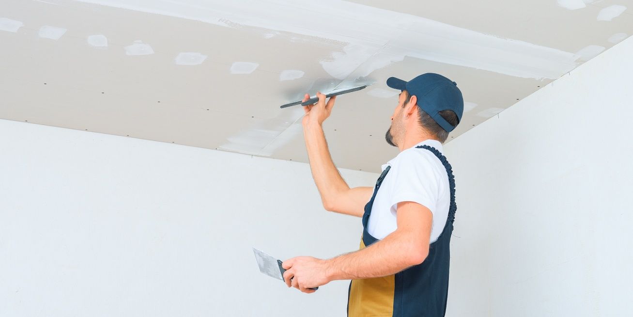 Man smoothing plaster on a white ceiling with a trowel, wearing a blue cap and overalls.