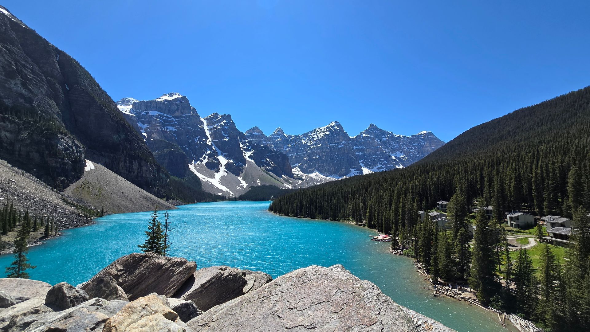 Turquoise alpine lake with rocky shore, pine forest, and snow-capped mountains under a clear blue sky