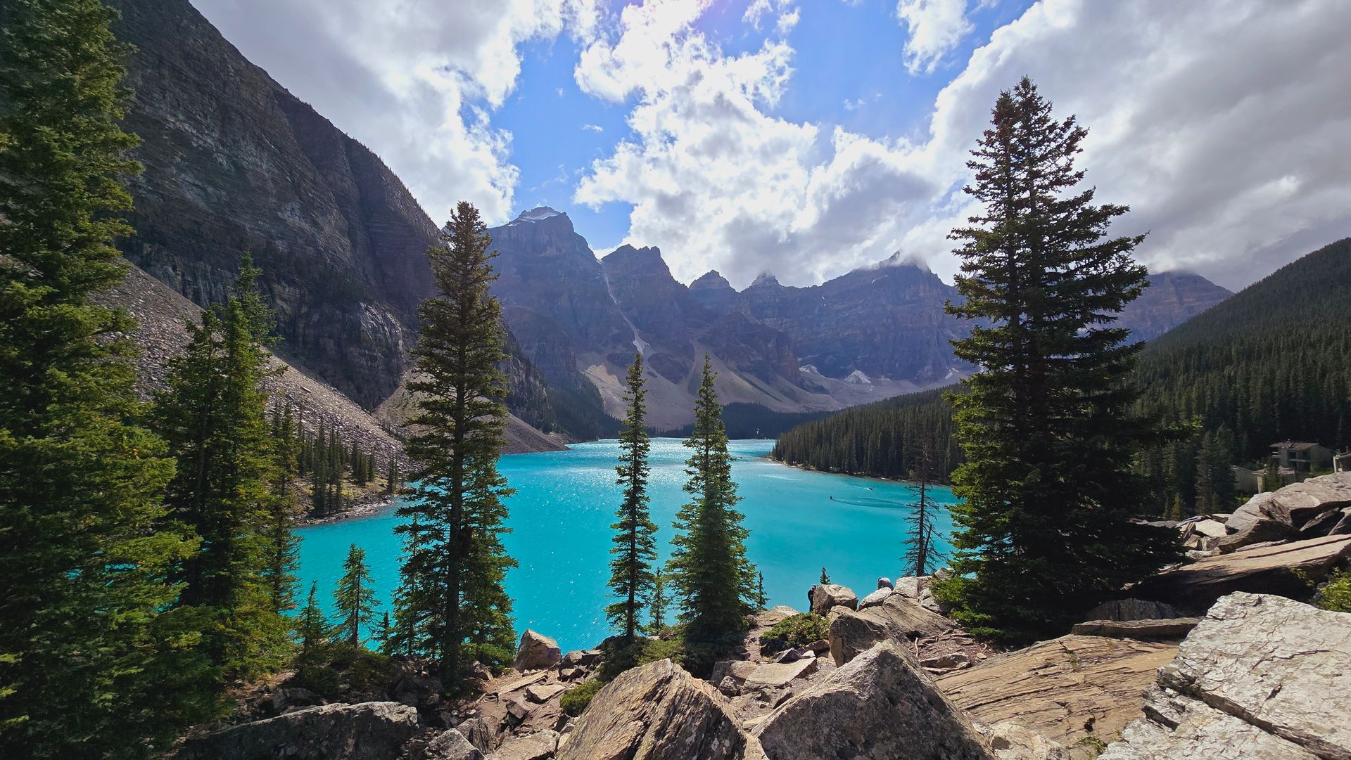 Moraine lake framed by pine trees and rocky peaks under a partly cloudy sky