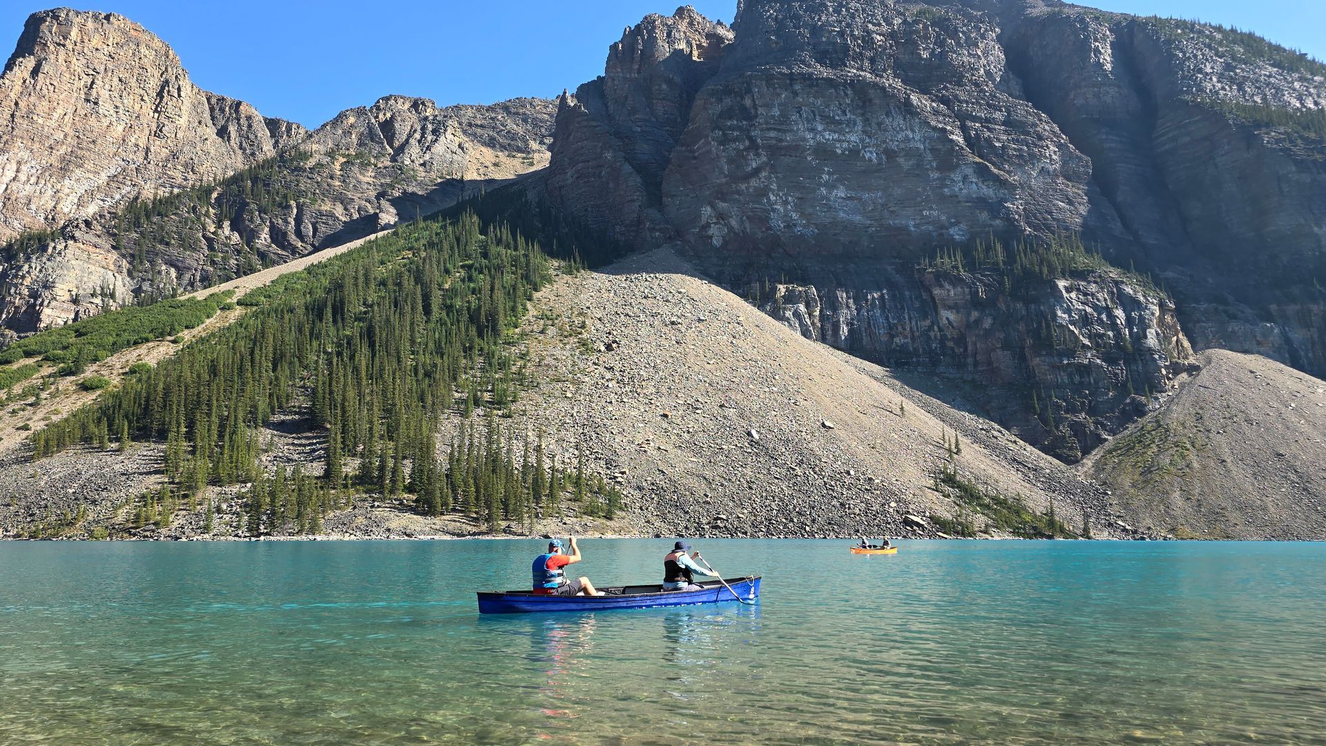 Two people paddle a blue kayak on a turquoise mountain lake beneath rocky cliffs.