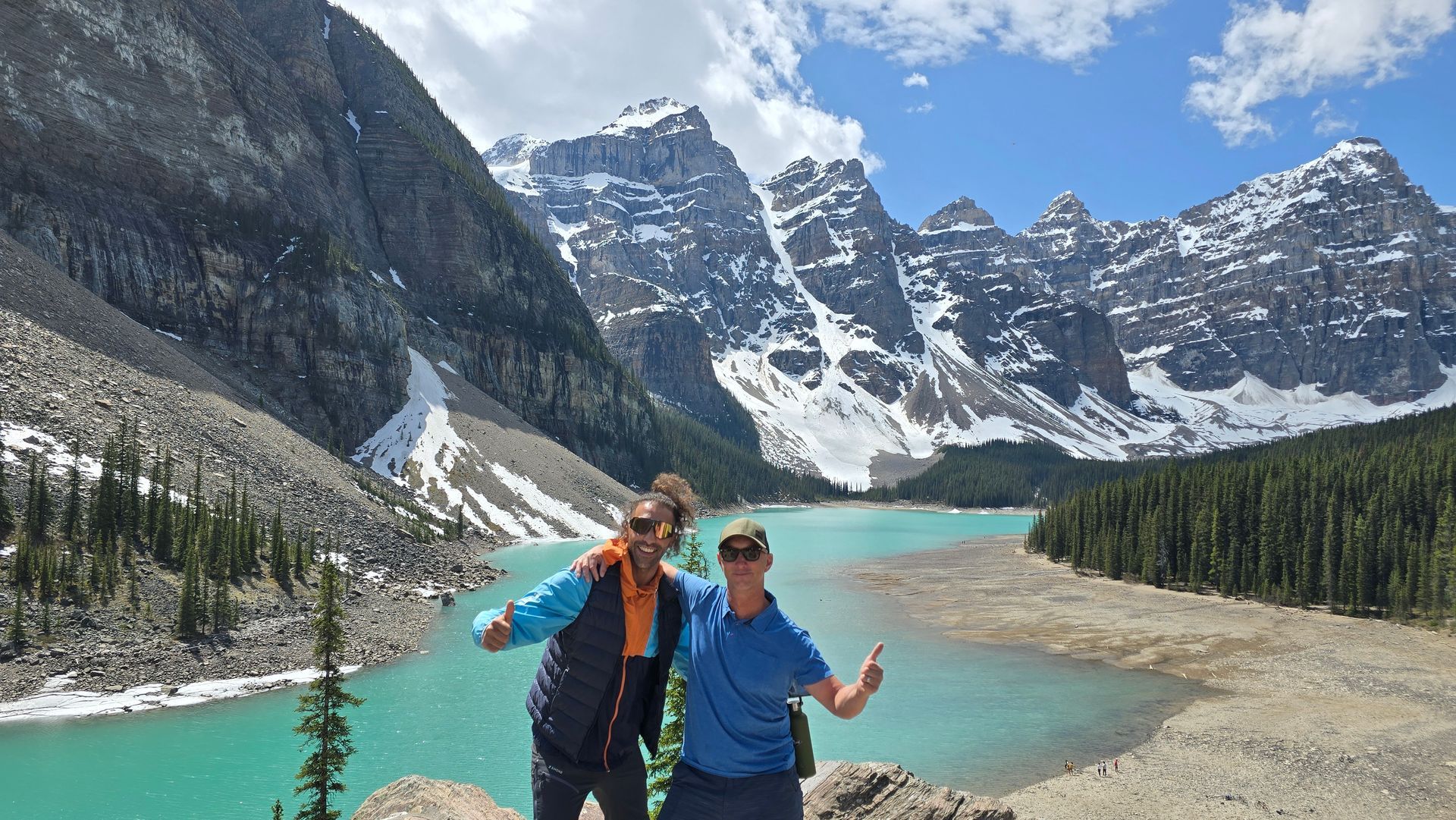 Two hikers posing by a turquoise mountain lake with snowy peaks and forested slopes.