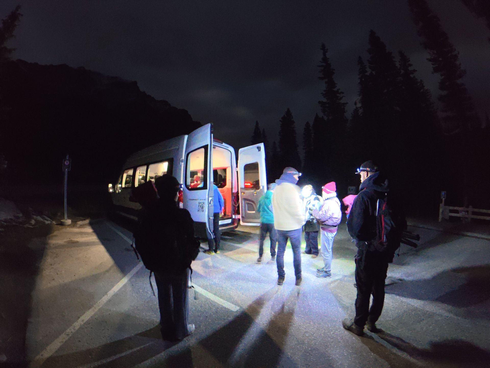 People boarding the shuttle at night on a dark road, lit by the bus and a flashlight.