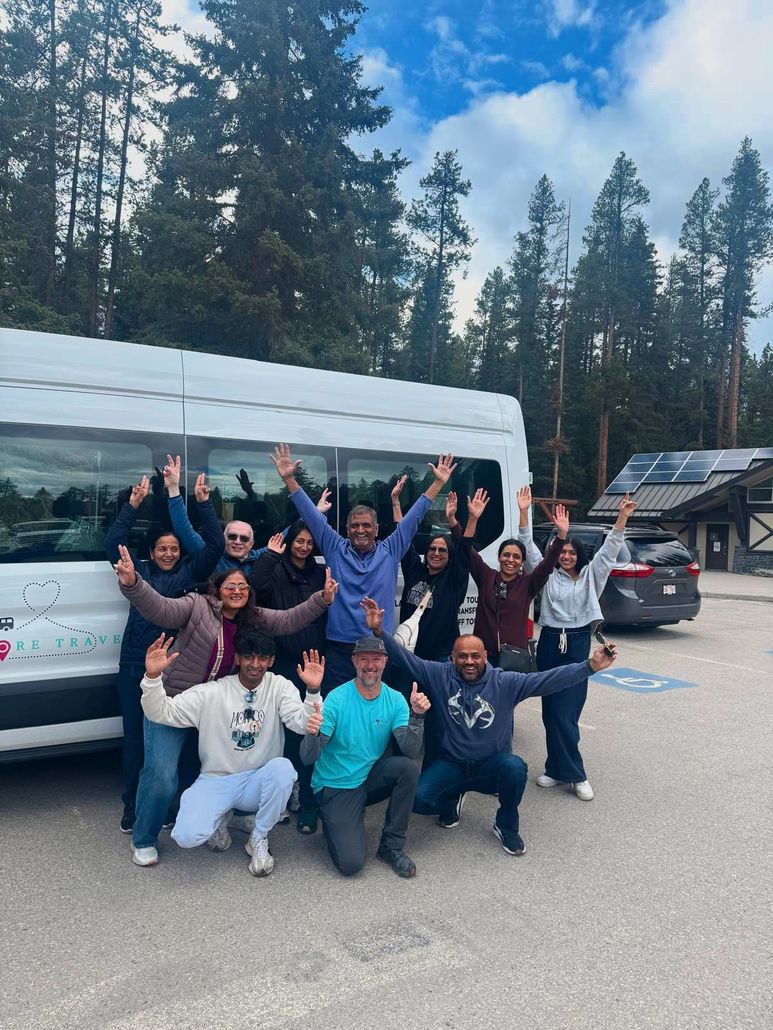 Group of people posing cheerfully in front of a white van in a forested parking area.