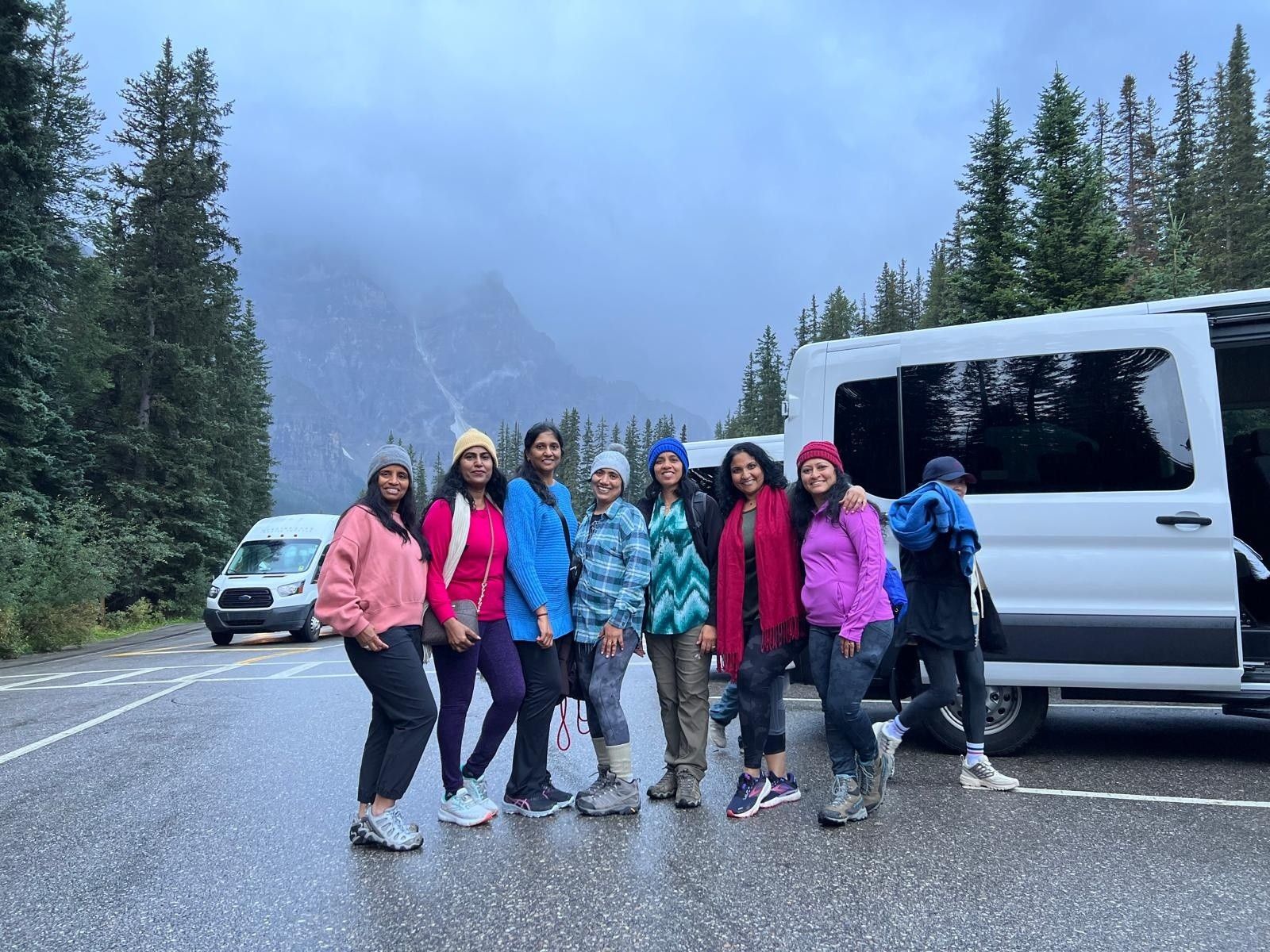 Group of hikers posing on a mountain road beside a white van, with snowy trees and misty hills behind them.