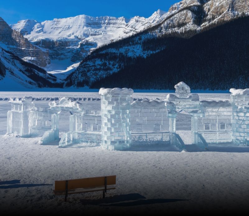 Ice sculptures on a frozen lake with snowy mountains in the background and a bench in front.