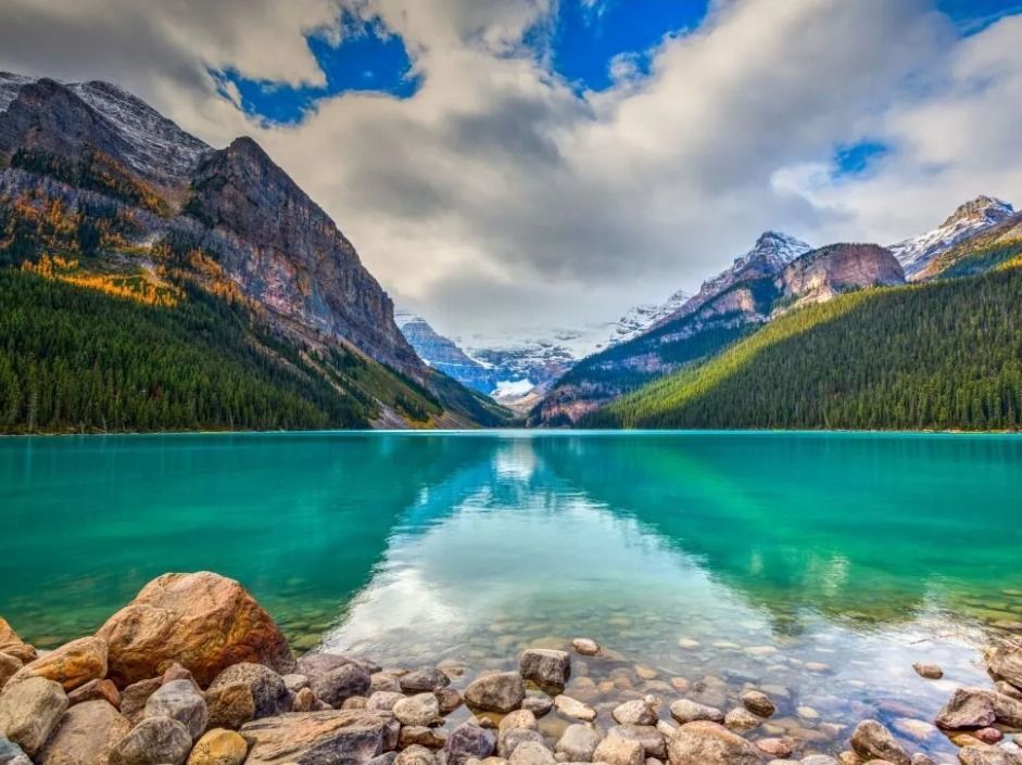 Turquoise mountain lake reflecting snowy peaks and cloudy sky, with rocky shore in foreground
