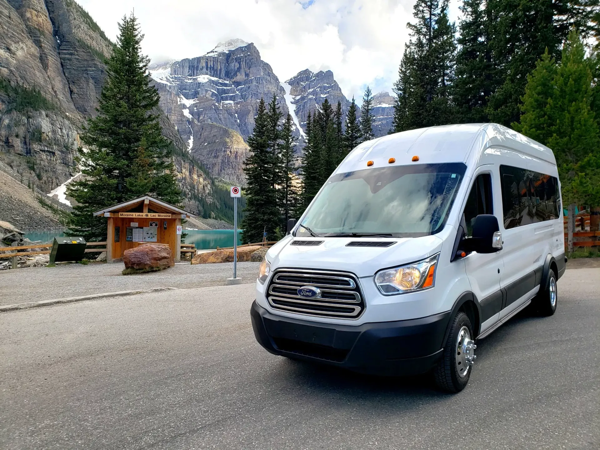 White shuttle van parked on a mountain road with pine trees and rocky peaks in the background