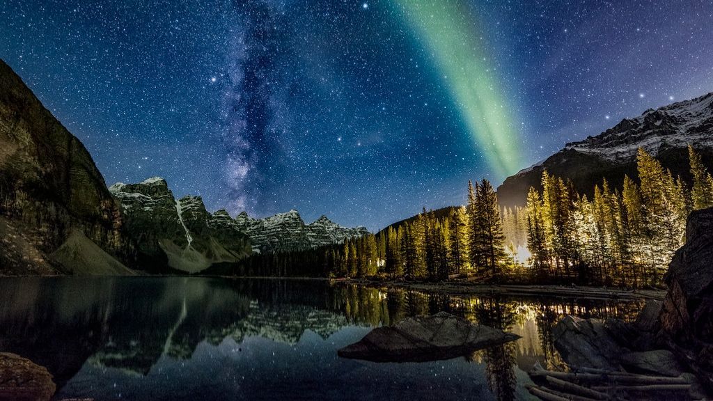 Starry night over a calm forest lake with glowing trees and green aurora above mountains