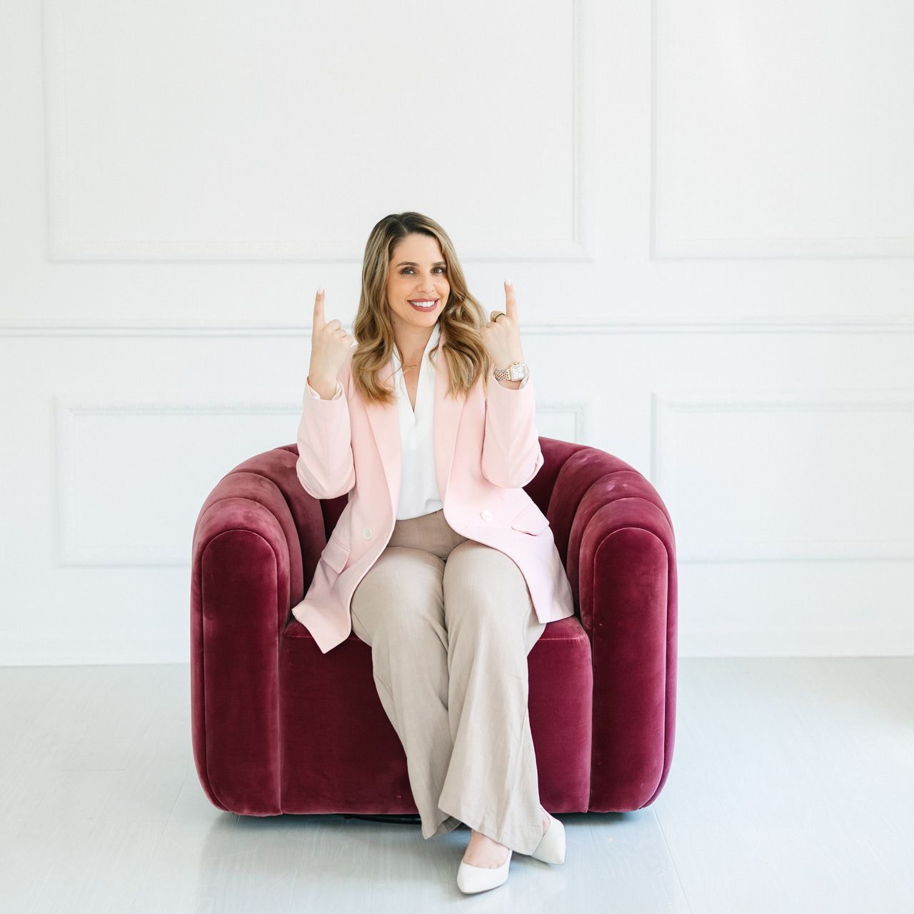 Woman in pink blazer and tan pants sitting in a red chair, making hand gestures in a white room.
