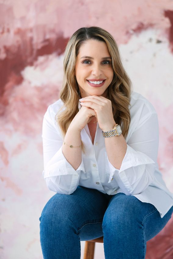 Woman smiling, wearing white shirt, blue jeans, arms crossed, sitting, pink art background.