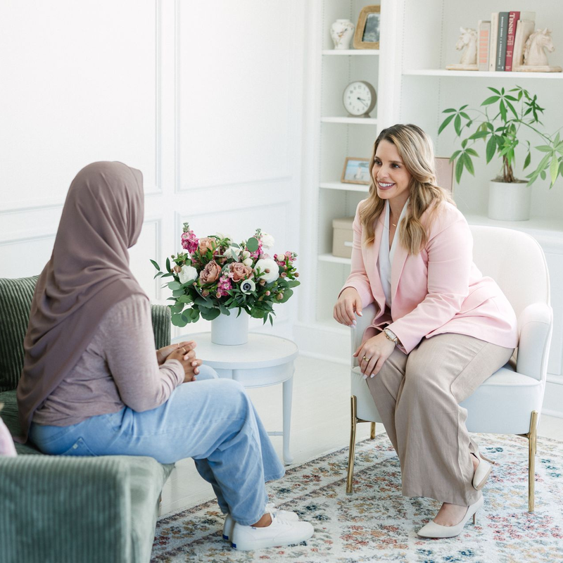Two women in a light, bright room having a conversation. One wears a hijab, the other a pink blazer, both smiling.