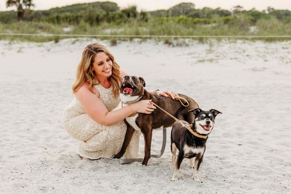 Woman on beach with two dogs; woman smiling, petting brown dog with black dog beside it.