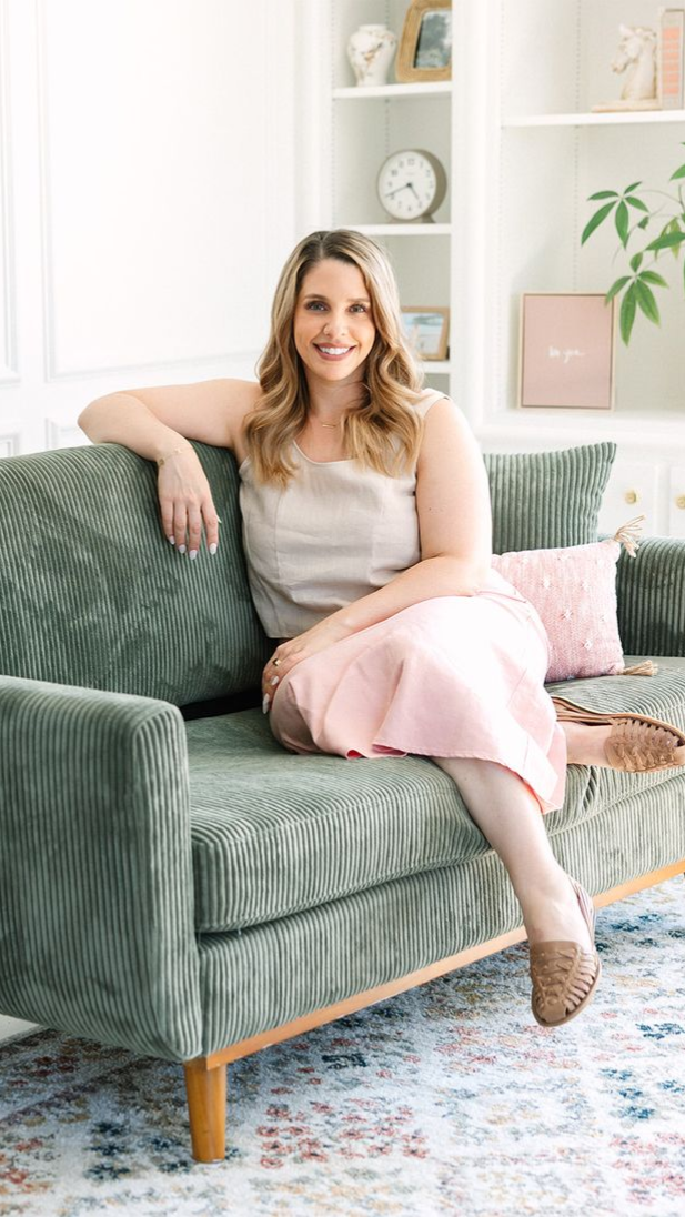 Woman in beige top and pink skirt sits on green couch; smiling indoors.