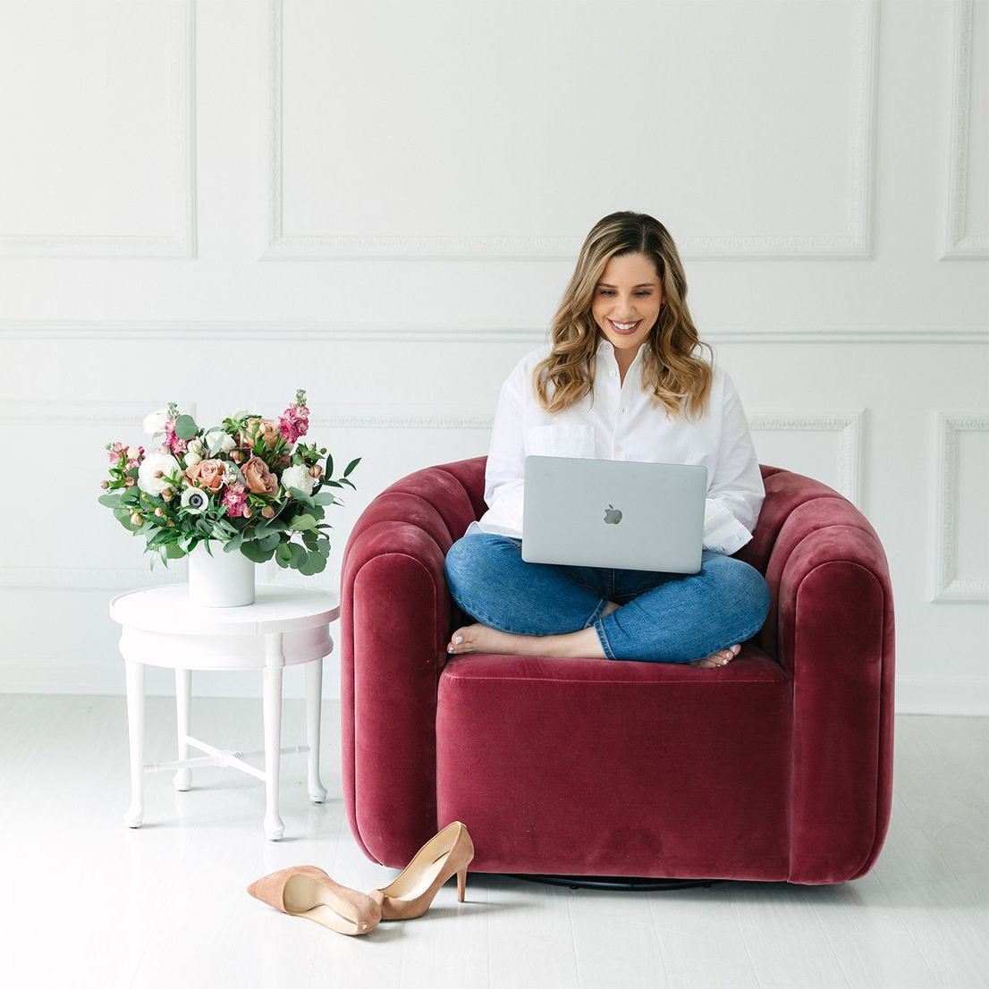Woman on red chair using laptop, legs crossed, smiles. Beige heels on floor, flowers and table to the left.