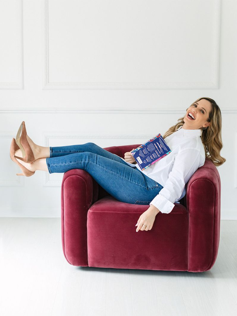 Woman in white shirt and jeans laughs with feet up on a burgundy chair, holding a book. White background.