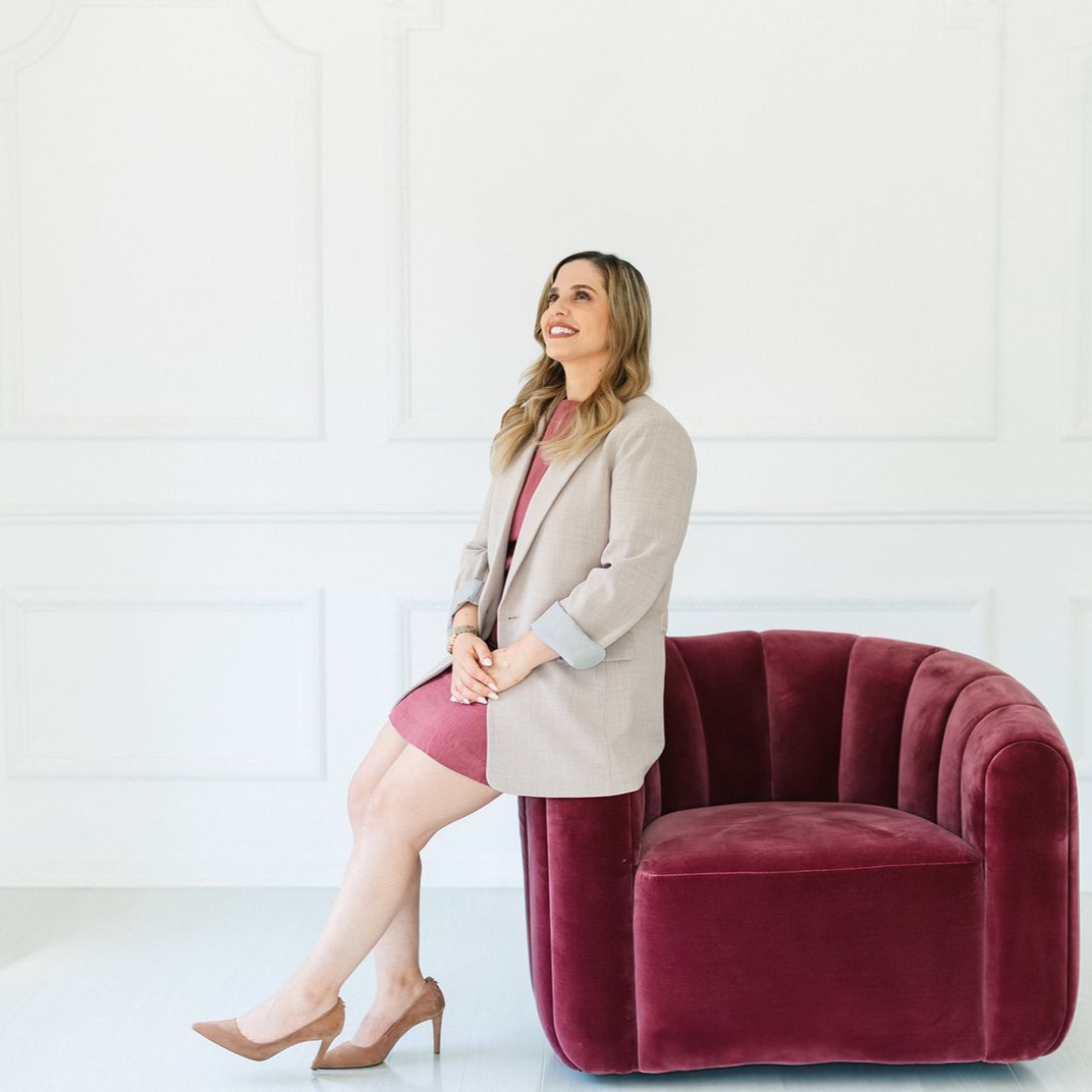 Woman in pink dress and beige blazer sitting in a burgundy chair, looking up.
