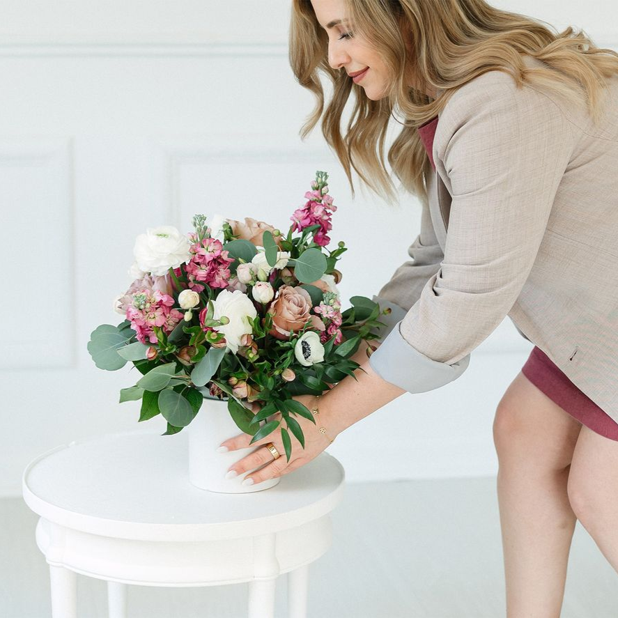 Woman arranging a flower arrangement in a white vase on a small white table.