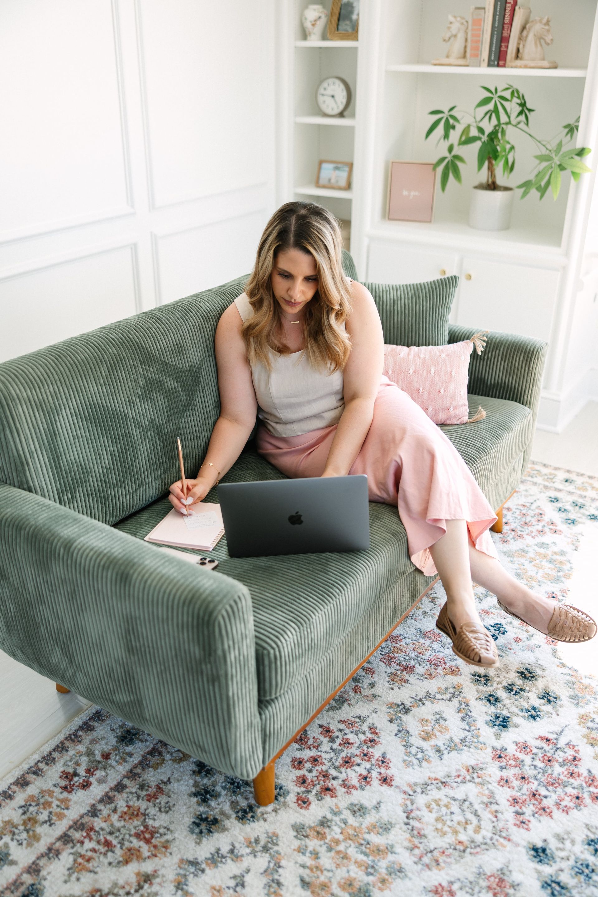 Woman working on laptop, taking notes on a green couch. Light-filled room.