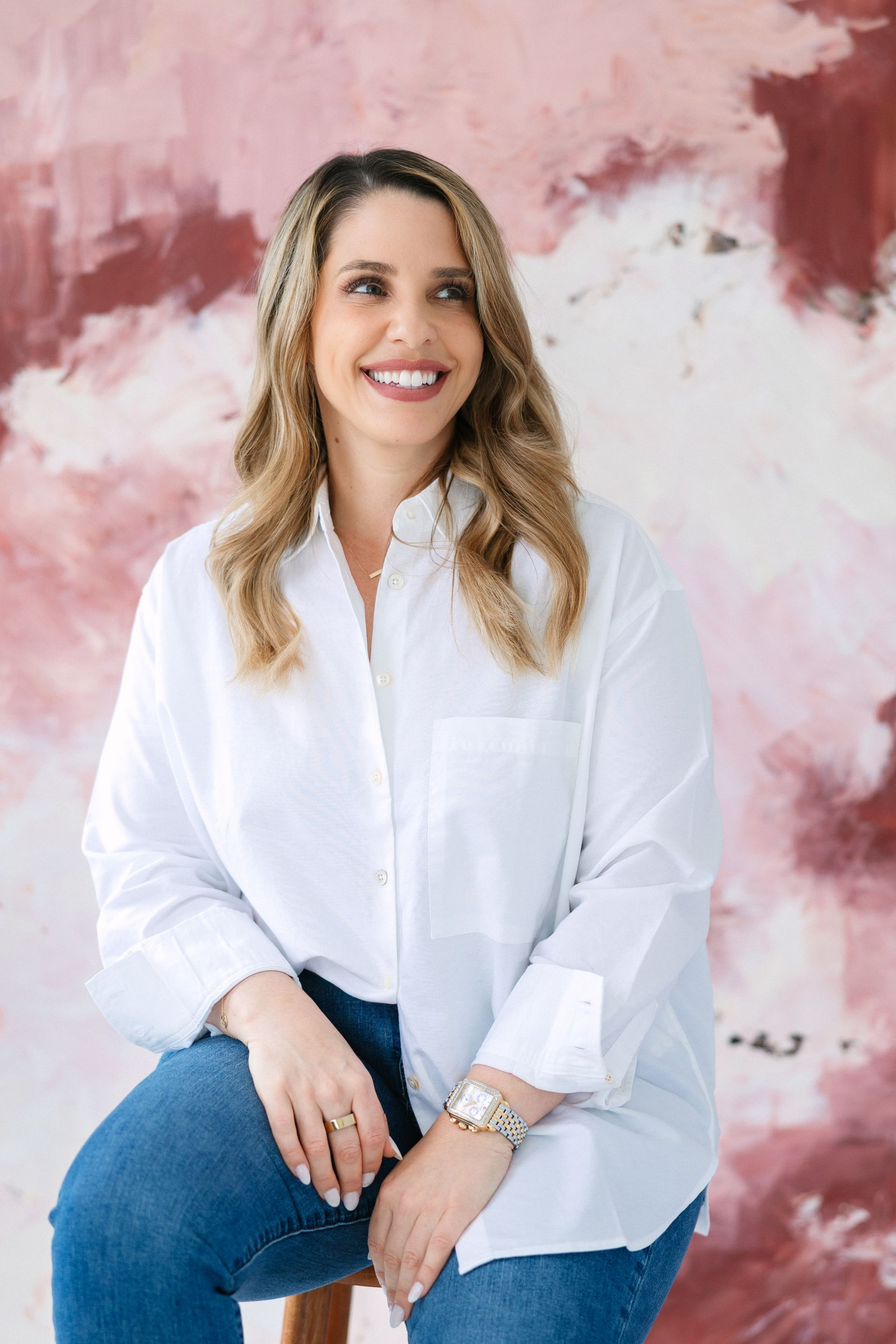 Woman with blonde hair smiles, wearing white shirt and jeans, sitting in front of abstract pink and red background.
