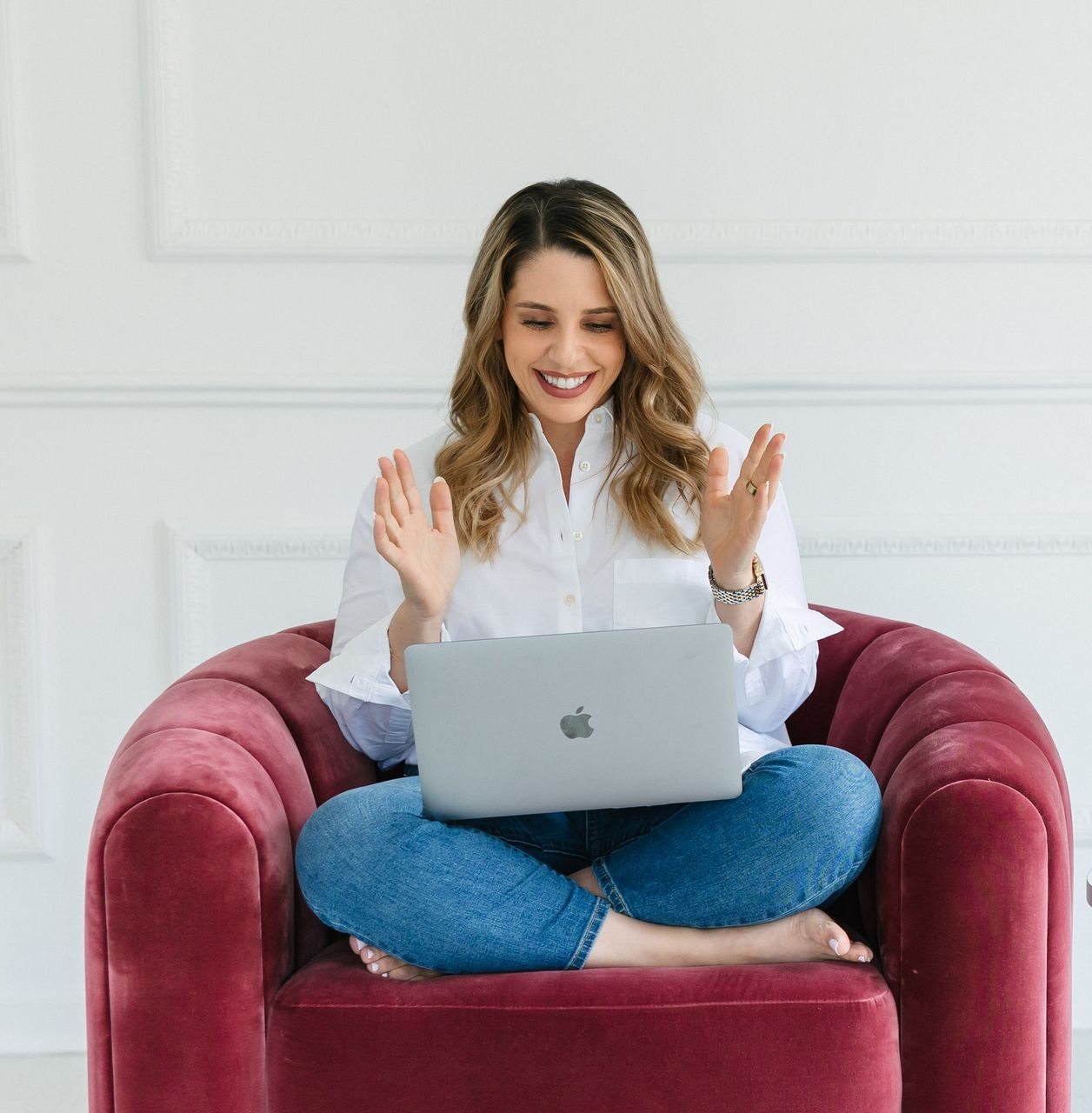 Woman with laptop, smiling and gesturing, sitting cross-legged in a red chair. White shirt, jeans, light room.