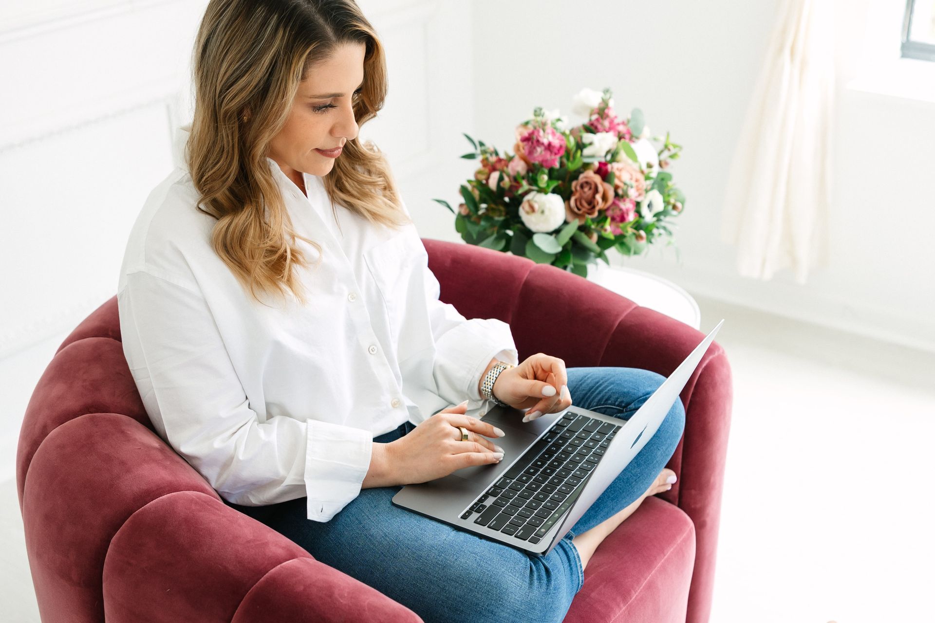 Woman in white shirt and jeans, using a laptop while sitting in a burgundy armchair. Bouquet of flowers. Bright room.