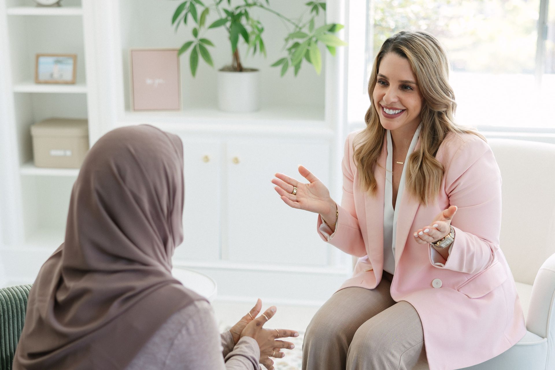 Woman in pink blazer gestures, smiling at a woman in a hijab in a bright, cozy setting.