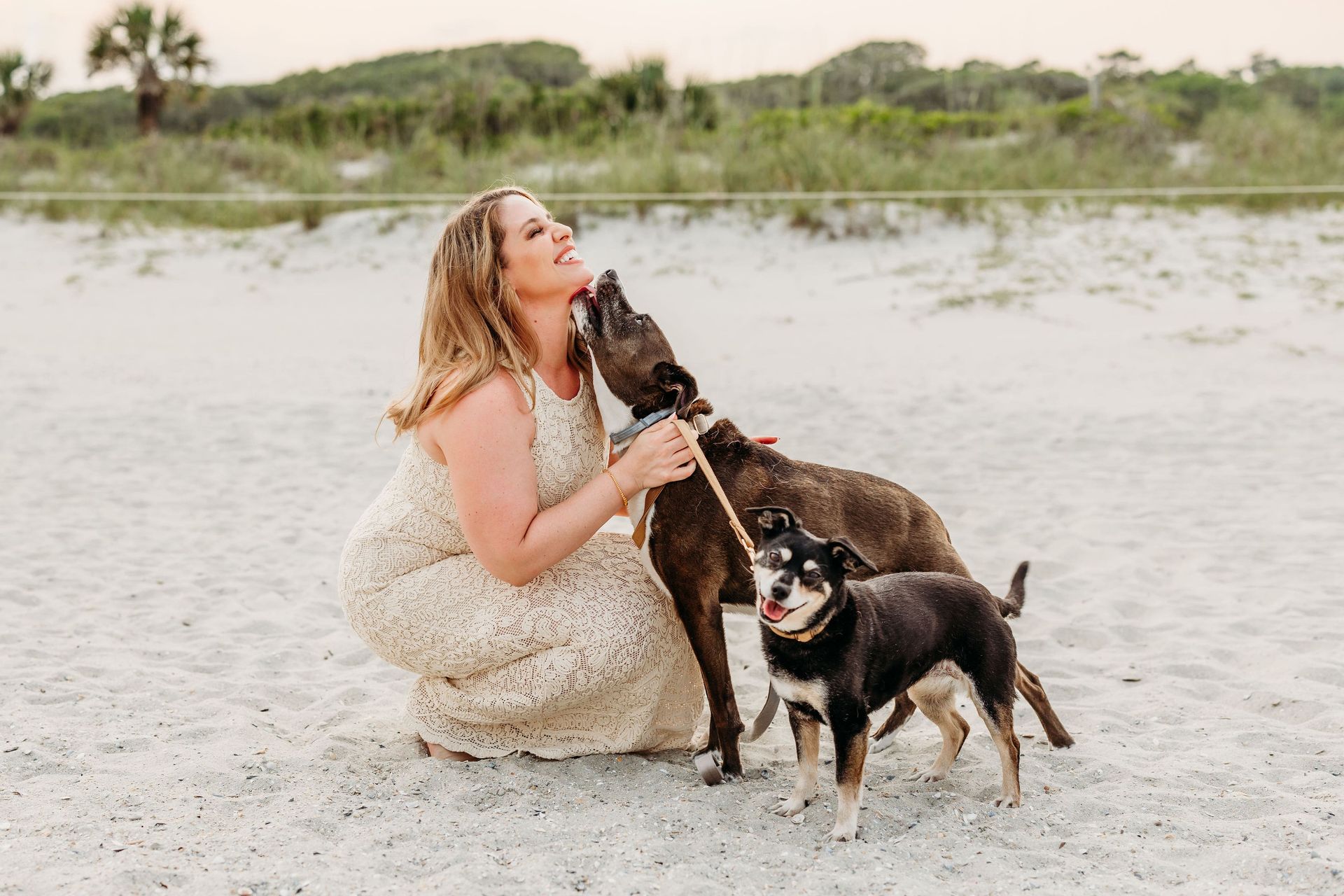 Woman on beach, smiles, kneels, pets two dogs. One licks her face.