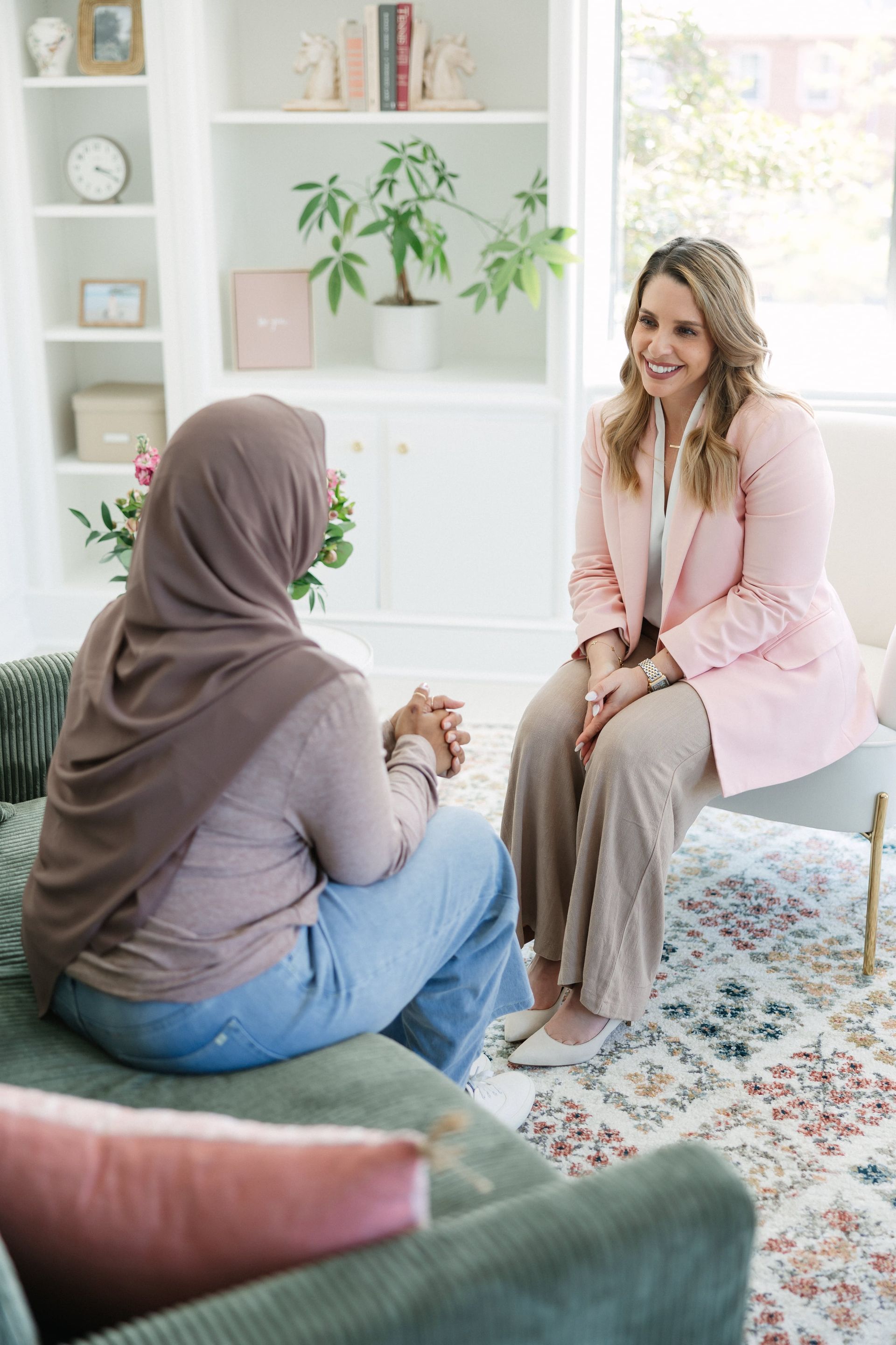 Woman in hijab on couch, talking to a smiling woman in pink blazer, in a well-lit room.