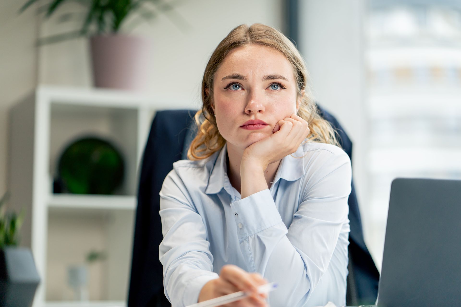 Woman in a light blue shirt, resting her chin on her hand, looking upward, at her desk in an office setting.