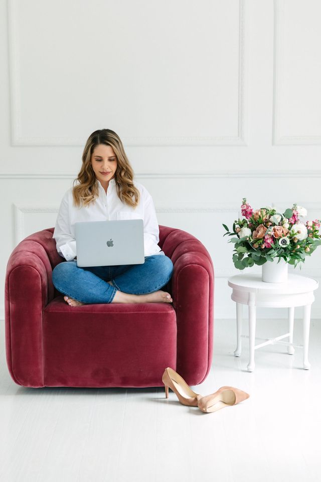 Woman in jeans and white shirt works on laptop, seated in red armchair, shoes nearby. White room, flowers on table.