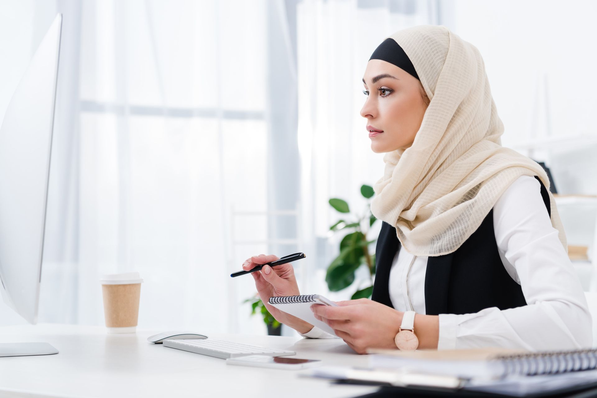 Woman in hijab working at a computer, taking notes, focused expression, light-filled office.