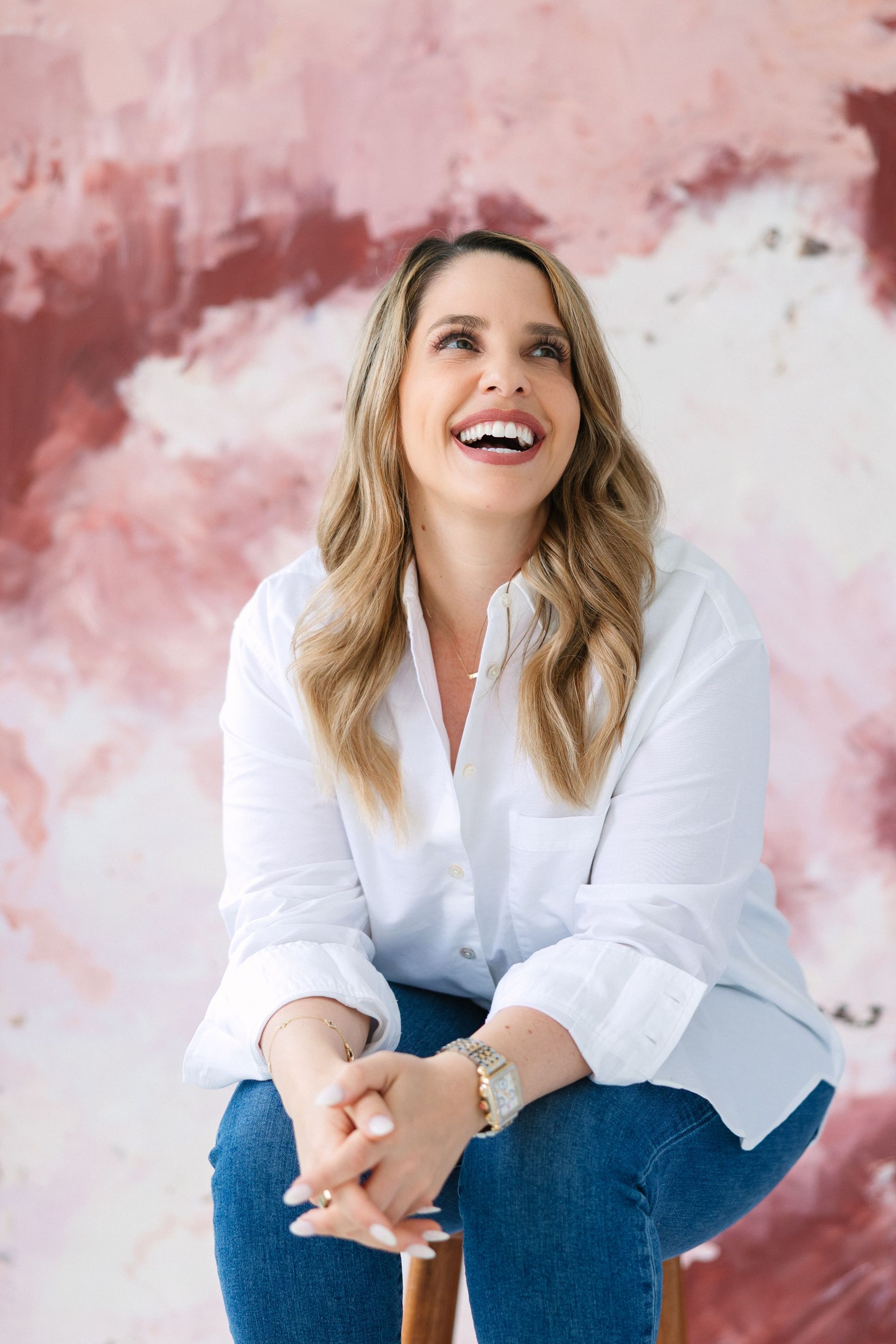 Woman laughing, wearing white shirt and jeans, seated, looking up with pink backdrop.