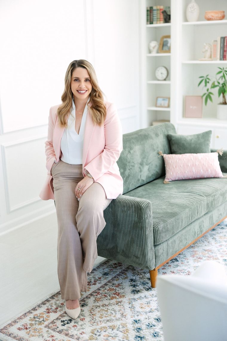 Woman in pink blazer leans on green couch in a well-lit room.
