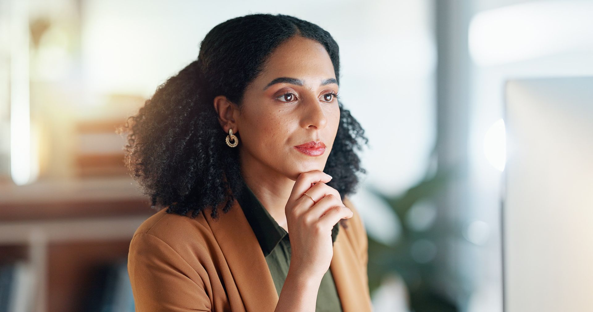 Woman in a brown blazer, hand on chin, thoughtfully looking at a computer screen in a modern office.