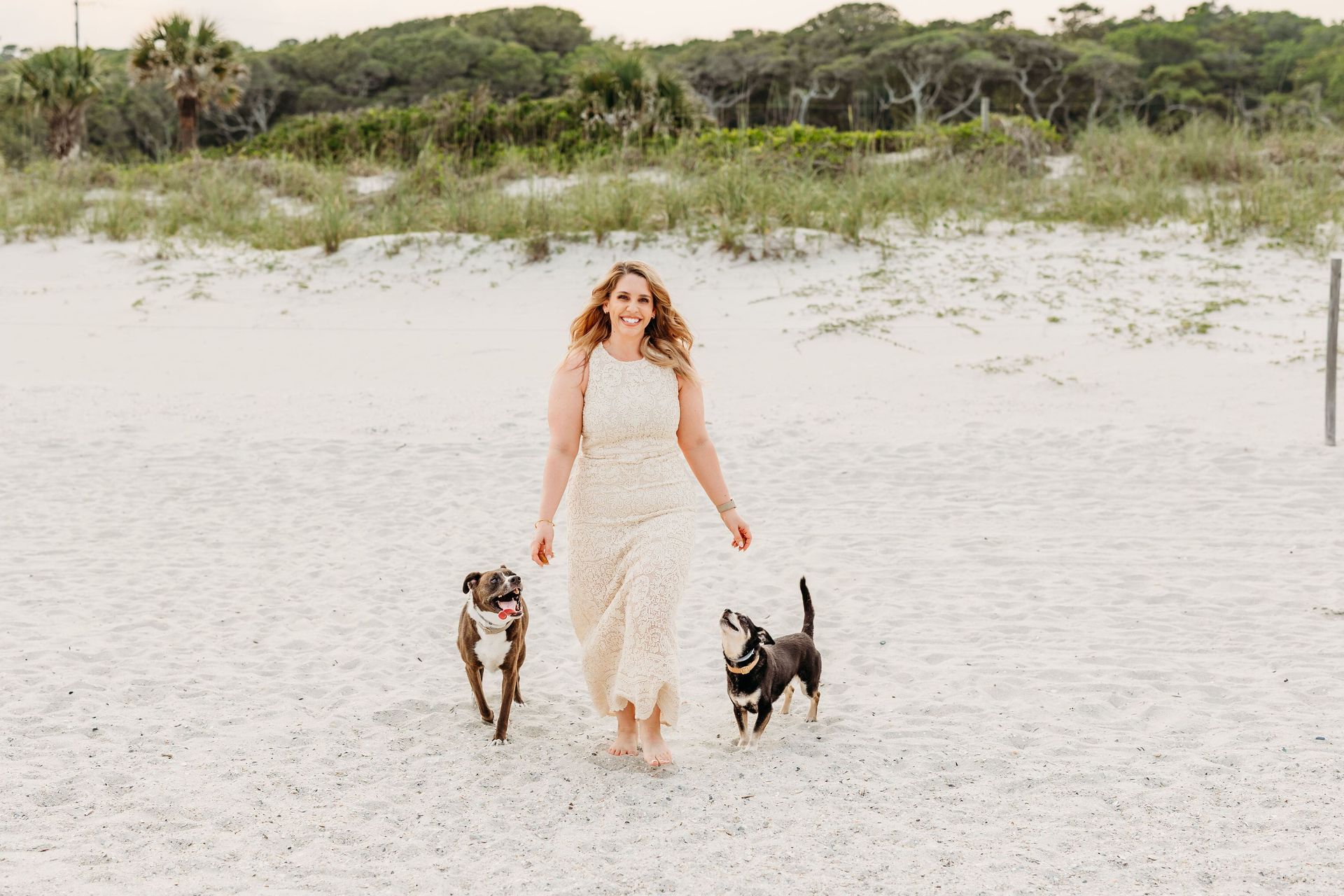 Woman walking with two dogs on a white sandy beach, wearing a light floral dress.