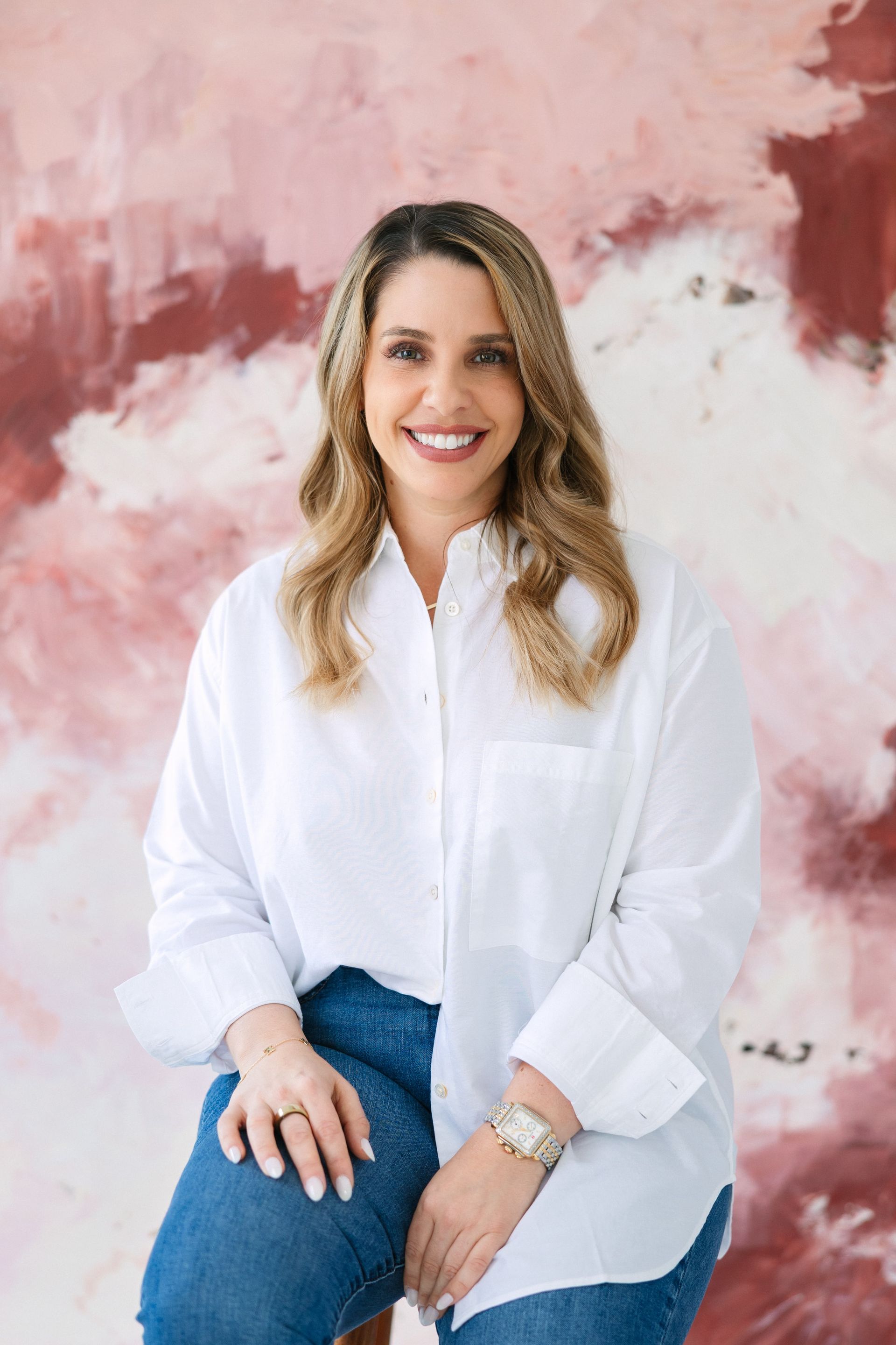 Woman in white shirt and jeans smiling, sitting, against pink-toned abstract background.