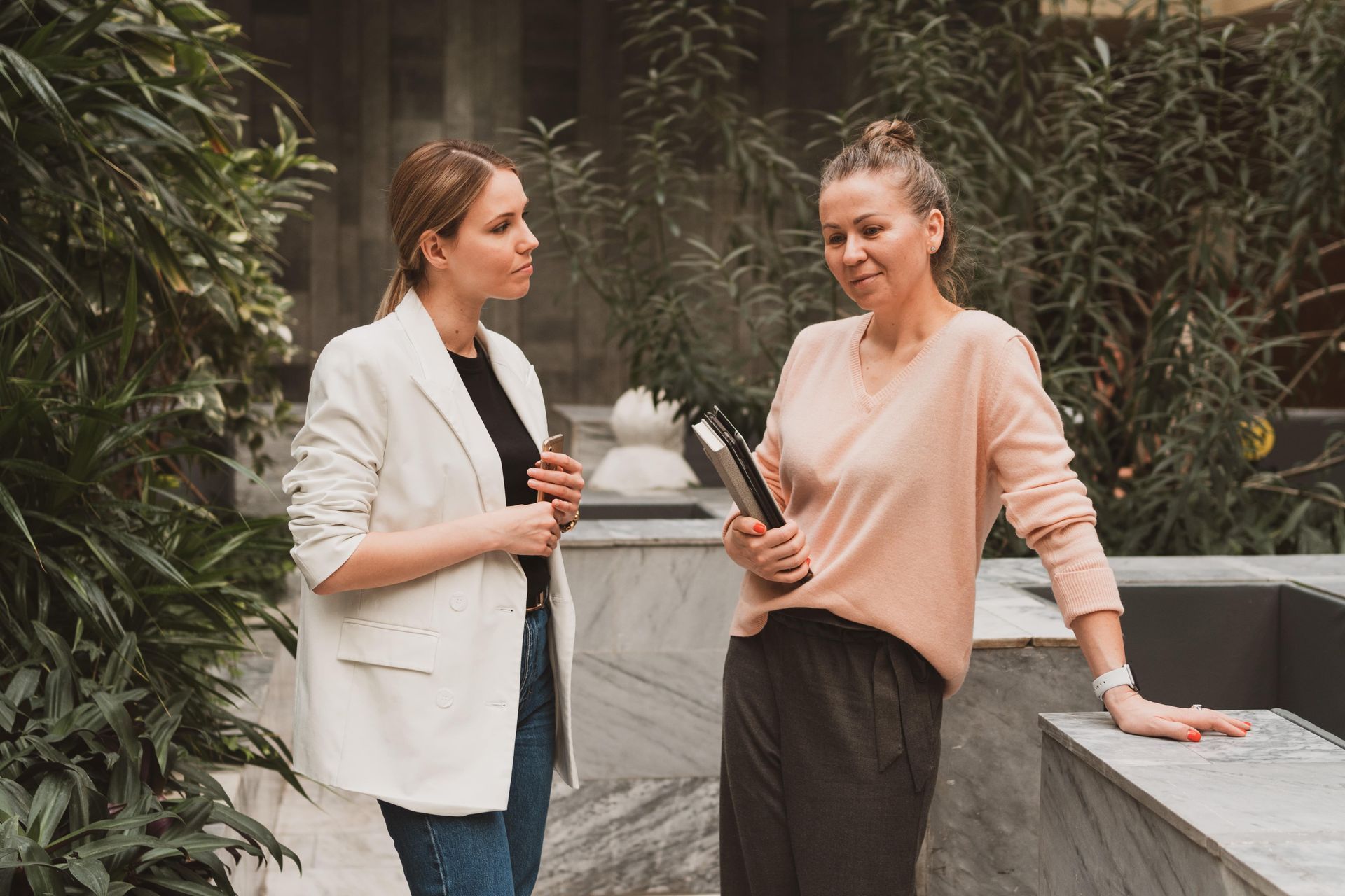 Two women talking near indoor plants; one in white blazer, the other in pink sweater holding a water bottle.