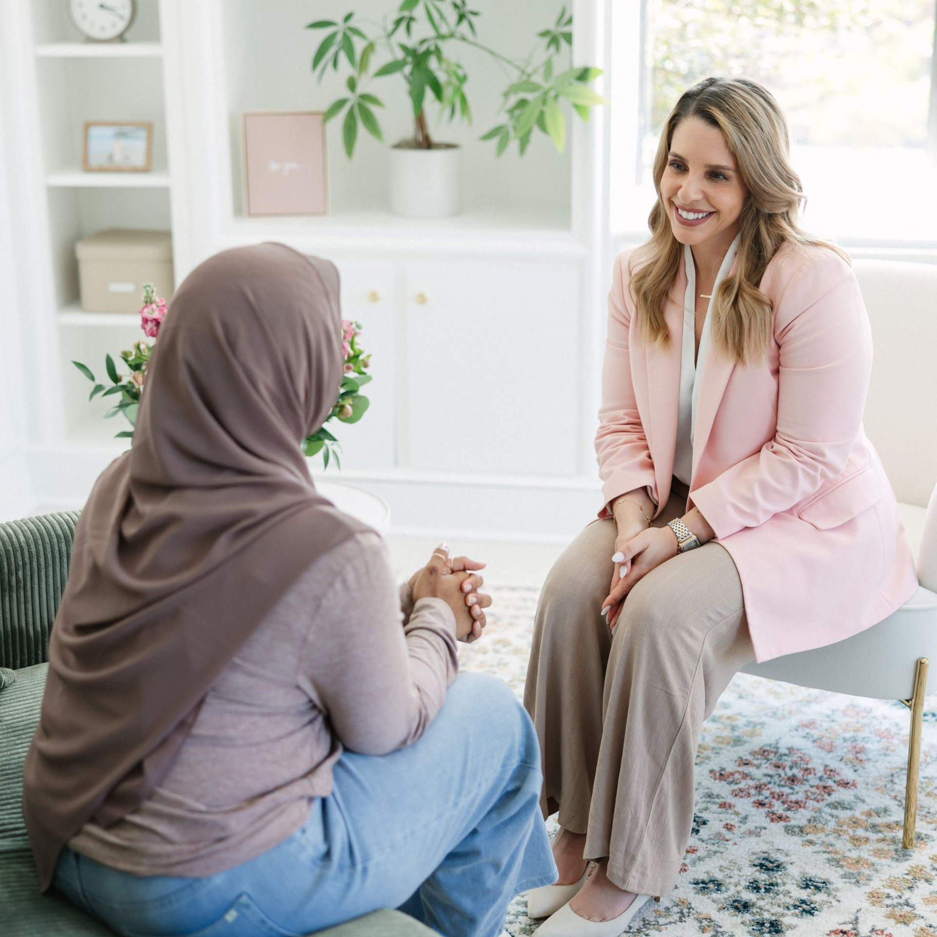 Woman in hijab consulting with a therapist in a bright, airy room; the therapist smiles, wearing a pink blazer.