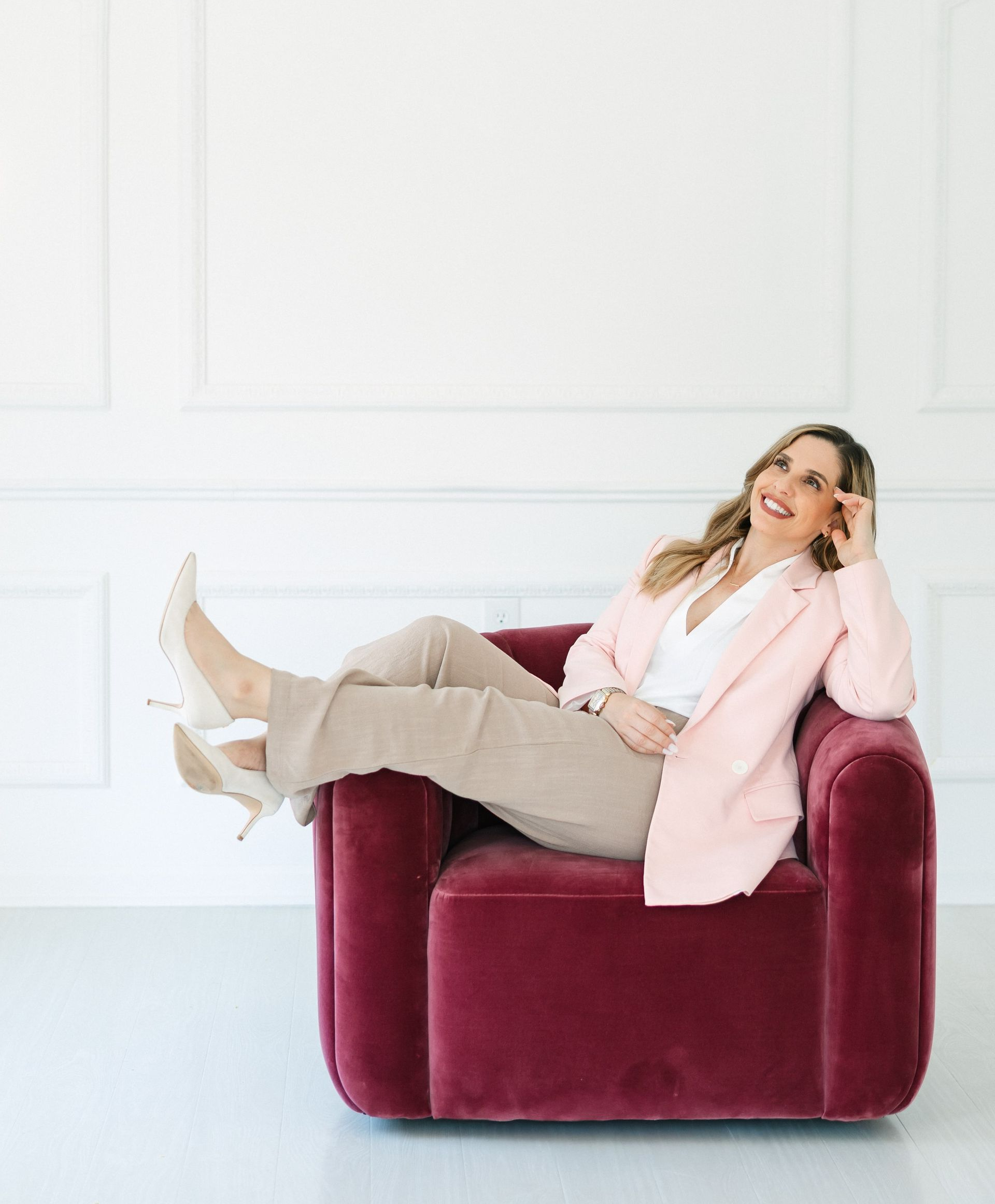 Woman in pink blazer and tan pants, lounging in a red armchair, smiling.