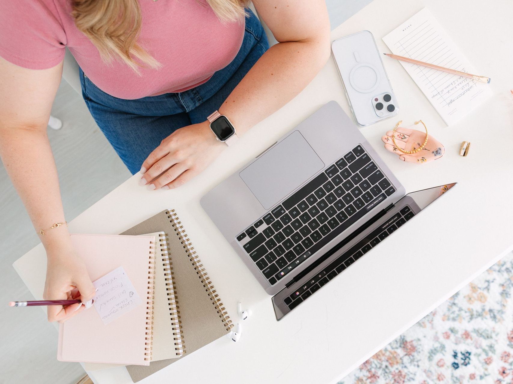 Woman in pink top at desk, writing in notebook. Laptop, phone, and accessories present.