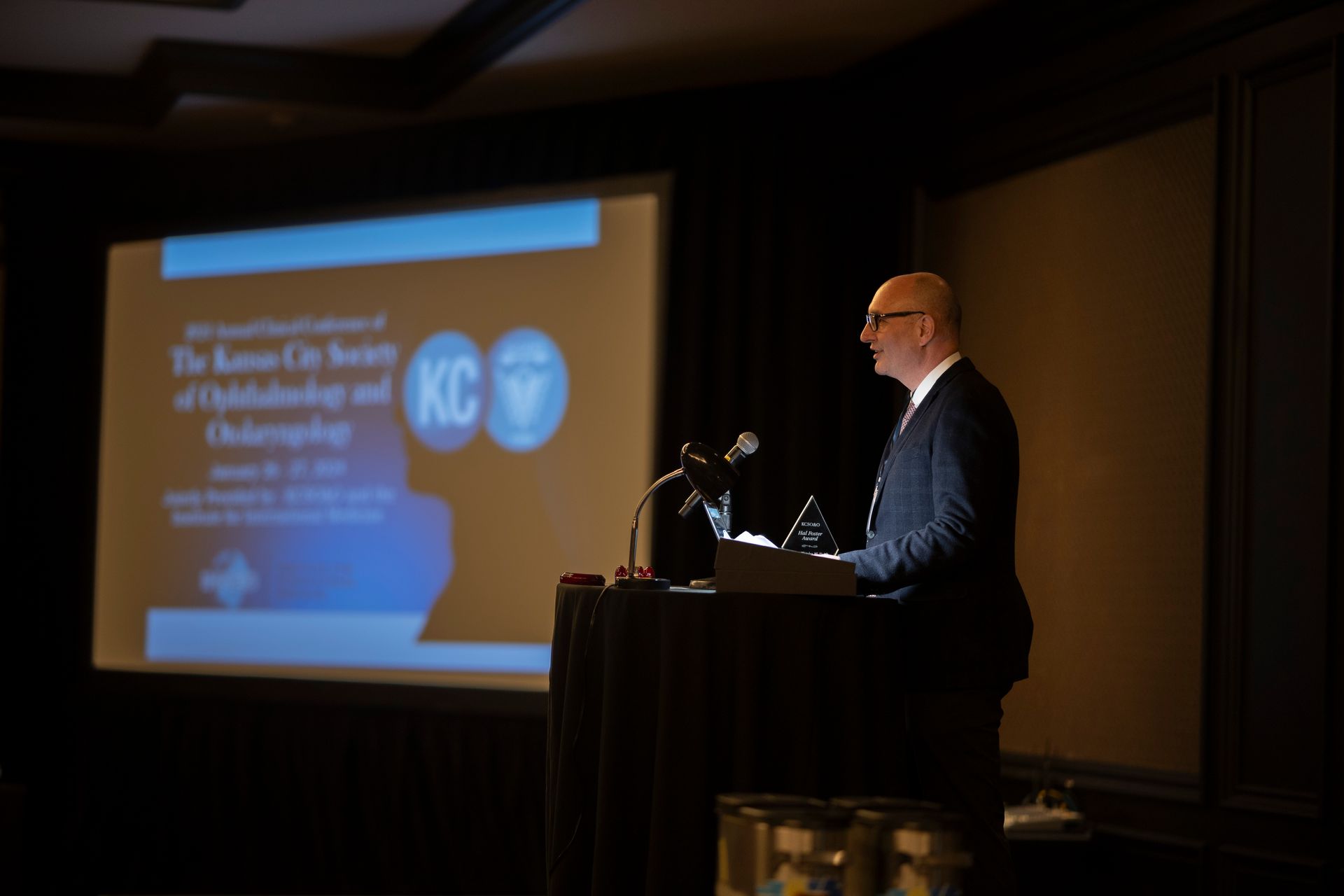 A man is giving a speech at a podium in front of a screen that says kc