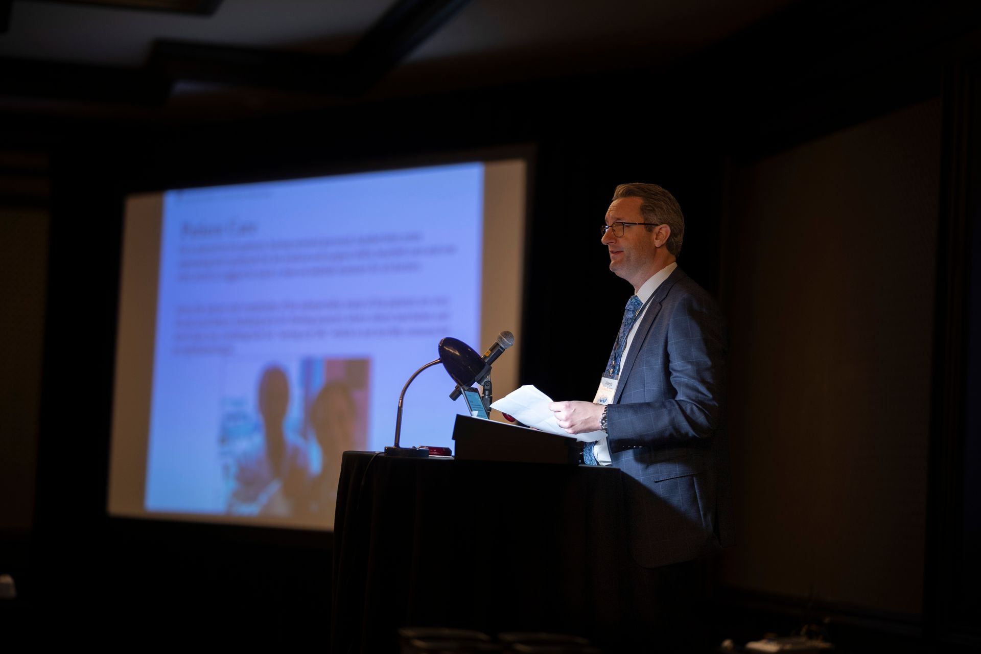 A man is giving a speech at a podium in front of a projection screen.