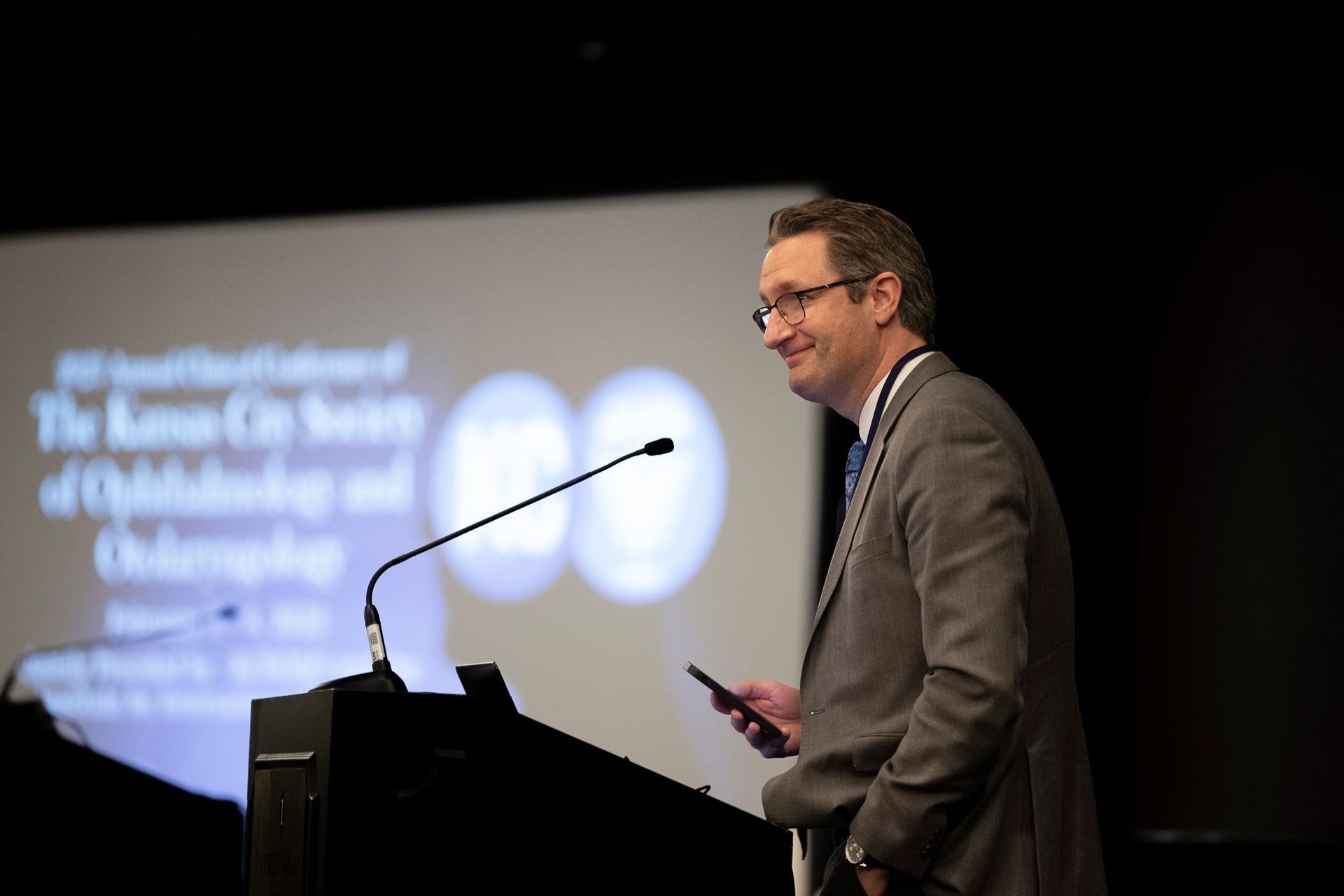 A man in a suit is standing at a podium holding a cell phone.